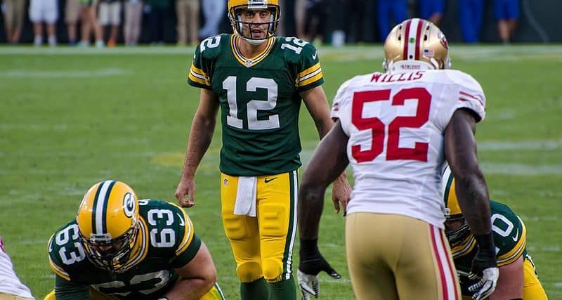A photo of Aaron Rodgers lining up to snap the ball against the 49ers. Rodgers is viewed straight on, while Patrick Willis is viewed from behind. Other Packers lineman are in the photo. - CC BY-SA