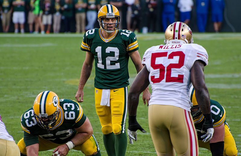 A photo of Aaron Rodgers lining up to snap the ball against the 49ers. Rodgers is viewed straight on, while Patrick Willis is viewed from behind. Other Packers lineman are in the photo. - CC BY-SA